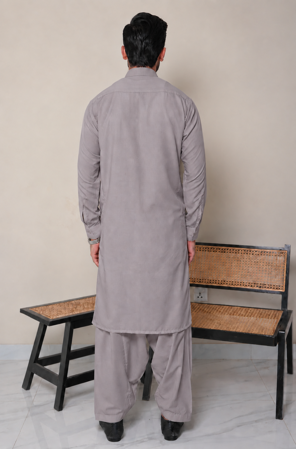 A man viewed from behind wearing a light gray traditional long kurta and matching trousers, standing in front of a beige wall with two wooden benches.