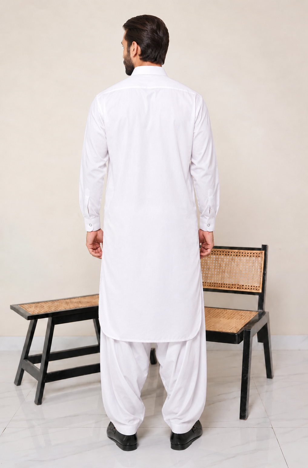 A man viewed from behind wearing a white long-sleeve kurta and white trousers, standing in front of a beige wall with a wooden bench behind him.