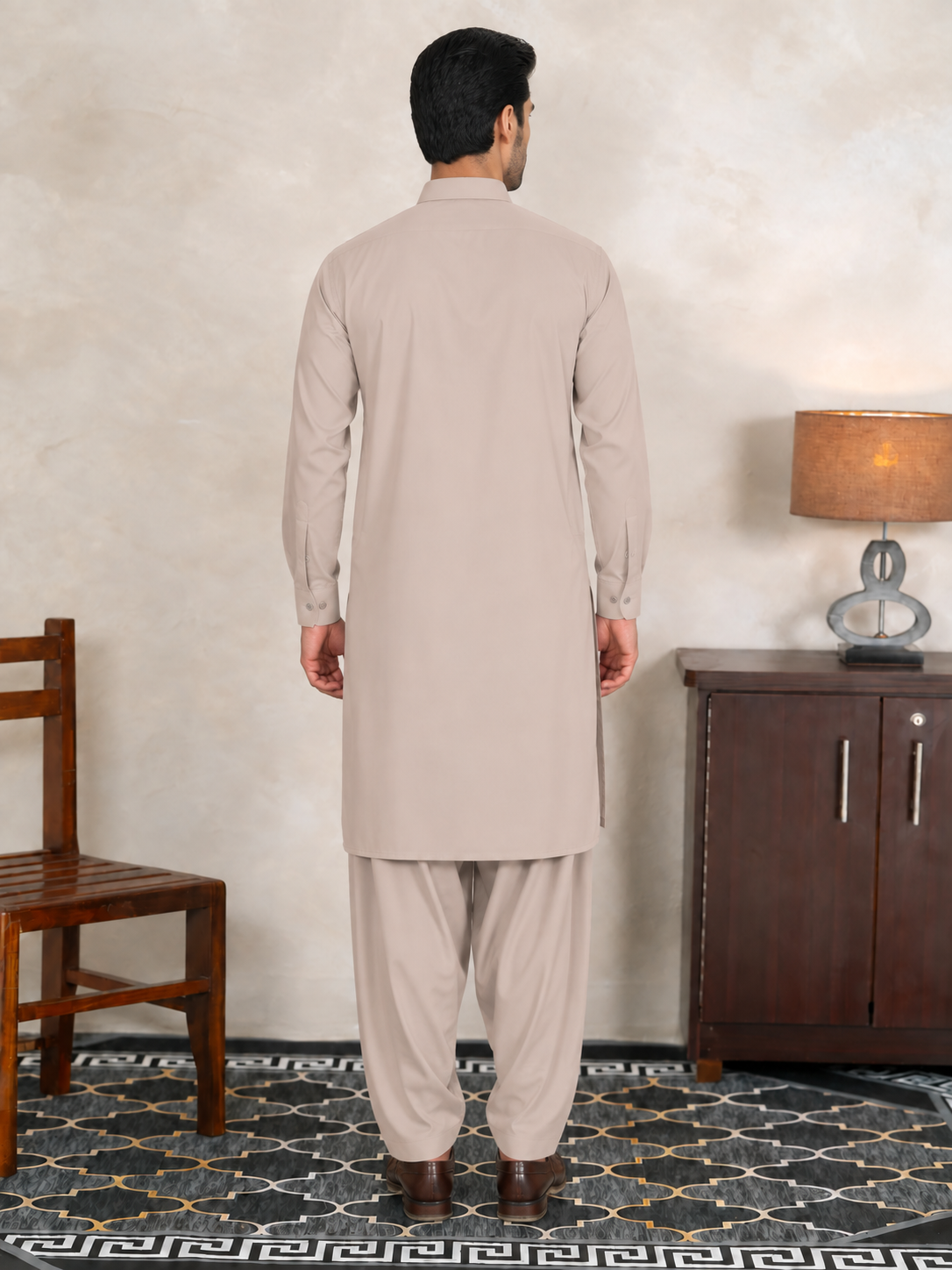 A man viewed from behind wearing a light beige long kurta and matching trousers, standing in a furnished room with a textured wall, chair, cabinet, and patterned rug.