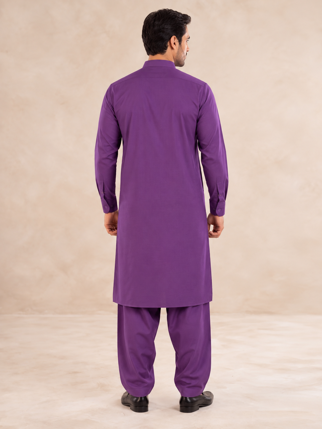 A man viewed from the back wearing a purple long kurta and matching purple trousers, standing against a plain beige studio backdrop.